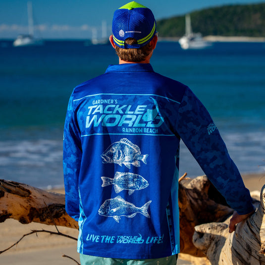 Person wearing a blue 'Tackle World' shirt on a beach with ocean and boats in the background